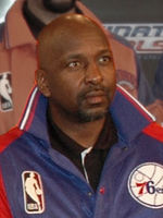 A man, wearing a black shirt and blue-red jacket with the logo NBA on it, is sitting on a chair while posing for a photo.