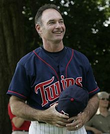 A man in a navy blue baseball jersey with "Twins" written across the chest holding a navy blue cap and smiling