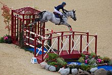 A gray horse, ridden by a woman, in mid-air over a red and white striped fence.