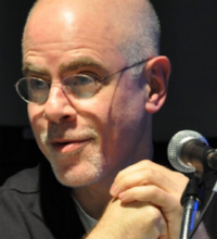 A photograph of a bald man wearing glasses and a black shirt. He is looking toward his right while a microphone is shown to the image's right.