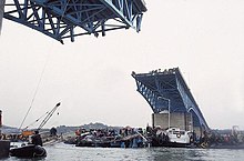 View of the missing bridge section from the Han River below the Seongsu Bridge. The day is cloudy, and there is a small crowd on the edge of the bridge. There are many boats and people crowding the collapsed section of the bridge, including a crane.