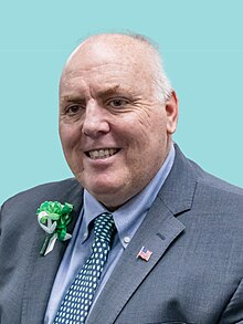 Flynn is shown in a front-facing bust-length photograph. He is wearing a grey suit jacket, a blue button-up shirt, and a blue tie tightly-dotted with a pattern depicting green four-leaf clovers in recognition of St. Patrick's Day. He is wearing a green corsage on his right lapel (shown on the lefthand side of the photograph), also in recognition of St. Patrick's Day. On his left lapel (which is shown on the righthand side of the photograph), he is wearing pin depicting the national flag of the United States. Flynn is an older caucasian male with shortly-cut grey hair which has partially-receded above his forehead.