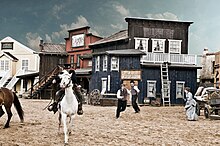 Background: Facades of Wild West themed buildings. A man is riding a horse and people are running in the background.