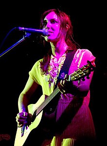 A twenty-nine-year-old woman is standing at a microphone. She is playing a six-string guitar with her left had on the fret board and her right at the strings. She wears over shoulder length red-brown hair and a light coloured dress. She stares out forwards.