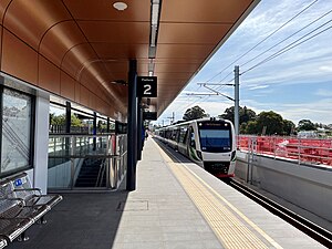 View down a station platform with curved shelter above with copper surface. An electric multiple unit train is on the tracks to the right.