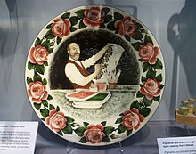 A painted plate, surrounded by cabbage roses, has a bearded man painting an umbrella stand with cabbage roses. At the bottom a caption reads "Karel Nekola 1857–1915".