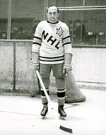 An ice hockey player stands on an ice rink. He is wearing a sweater that has the letters "NHL" in a downward diagonal with a large star on his left shoulder.