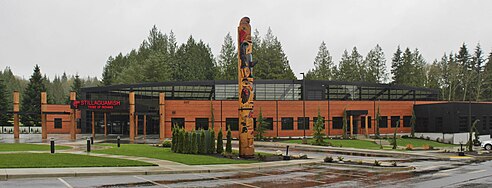 A large cedar building with a glass ceiling. There is a large totem pole near the entrance, with a parking lot in the foreground