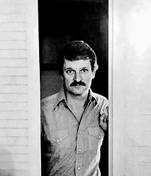 A black and white photo of a man with shaggy black hair and a moustache standing in a doorway.