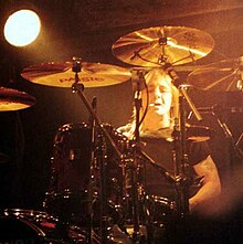 Rudd, aged 42, mostly obscured by his kit. He smokes a cigarette, has shoulder-length hair and wears a dark T-shirt. He has a tattoo on his upper left arm, right arm obscured.