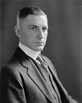 A man in his late twenties with dark, short, slicked-back hair and wearing a black coat and tie and white shirt