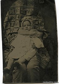 Toddler in a white coat and knitted hat and scarf sat on a pair of trousered knees. The head, torso and arms of the second person are hidden under fabric.
