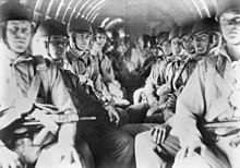 Men with helmets sit in an aircraft with weapons held across their chests, strapped into parachutes