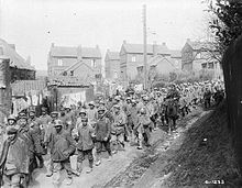 a large crowd of German soldiers walking along a small road under the supervision of soldiers wearing British equipment on horseback. Multiple houses can be seen in the background.