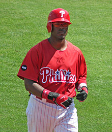 A dark-skinned man in a red baseball jersey, white baseball pants with red pinstripes, and a red batting helmet walks on a baseball field while taking off his black and red batting gloves.