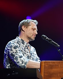 Andrej Šeban wearing patterned shirt, sitting down in front of microphone, with hands partly concealed