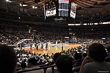A basketball arena viewed from far away in the stands during a game. The overhead scoreboard and entire court are in view while a player at the near free throw line is shooting free throws.