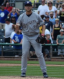 A man in a gray baseball uniform with "Brewers" across the chest and a navy cap standing on a pitcher's mound with a ball in one hand
