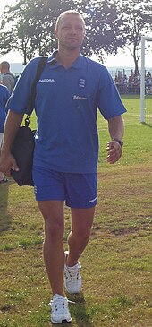 A young white man of athletic build wearing a blue sports shirt and shorts and carrying a kitbag walks across sunlit grass. In the background are trees and the edge of a football goal.