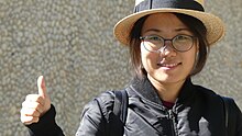 A close-up of a young Chinese woman wearing a black jacket, a straw hat and round glasses smiling and giving a "thumbs up" to the camera