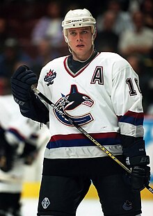 A hockey player on the ice before spectators. He wears a white jersey with a stylized C logo, and his youthful face has a serious expression.