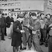 Six soldiers on the right and civilians on the left. Two soldiers at the front talking to a civilian female dress in black