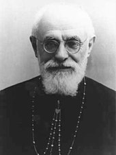 A bearded and bespectacled elderly man wearing a pectoral cross around his neck faces forward.