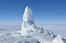 A photo showing an ice-covered landscape with a large tower of ice in the centre and a human for scale