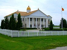 A photograph of a two-storey building. The roof is topped with a large cupola and three visible kalashes. Golden letters on the pediment say "CAPITAL OF THE GLOBAL COUNTRY OF WORLD PEACE". There is a circular driveway with a flagpole bearing the GCWP's sunburst flag. The building and drive are surrounded by a white picket fence, green lawn, and shrubbery.