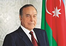 Picture of Heydar Aliyev, a smiley man in his 70s, standing next to the flag of the Republic of Azerbaijan.