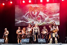 A photo of seven people on a stage, all dressed as Vikings, holding instruments like microphones and guitars, with drums in the back. There is a poster of their album Emblas Saga in the background.