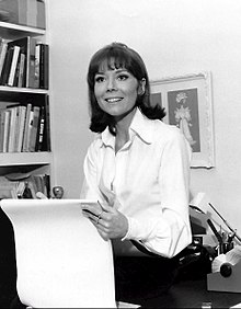 Diana Rigg sitting on a desk, smiling, holding a large pad of paper
