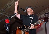 Country music singer Toby Keith singing into a microphone, holding the neck of a guitar with his left hand while his right hand points upward.