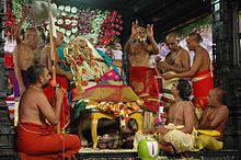 A priest holds up a three-disc gold mangala sutra to the audience while others look at the idols.