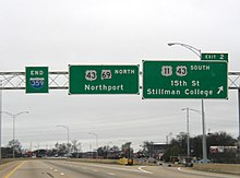 Three green signs are located above an elevated portion of roadway with no traffic visible on a cloudy day.