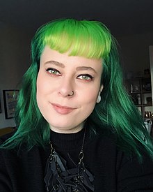 A white person with long light green hair with bangs. She is wearing a green dress with long necklace. She is also wearing a small black nosering on left side, and small white earrings. She is looking at the camera, with slight smile. In the background there is a shelf with decorative items on the left, and a poster on the wall, on the right.