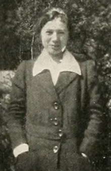 A young white woman, standing outdoors, wearing a white collared blouse and a dark jacket; her hands are in her pockets