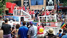 Multiple people on a parade float passing a street lined with people. At the back of the float, left of the image, is a large red T representing the network logo. Several banners promoting Telemundo Chicago programming and diversity initiatives are attached to the skirt of the float.
