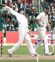A Black bearded man wearing a black turban in his bowling action. The stumps, off-strike batsman, field, boundary ropes and the spectators can be seen in the background.