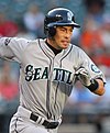 A man in a gray baseball uniform running to the right. The uniform reads "Seattle" across the front, and has a sleeve patch with a teal and silver compass surrounded by "Seattle Mariners". The man is wearing a dark batting helmet.