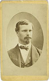 A man in a suit and bow-tie is shown from the chest up, facing slightly right of the camera.