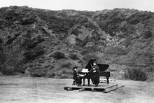 On a platform in a field, against a hillside, one woman plays a piano while a woman singer stands nearby