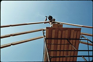 Pipe staging is very common in the U.S. Welded sections stack on top of each other and braced with cross braces, workers stand on planks or aluminum platforms, 1975