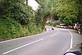 Racing motorcycle negotiating the entrance to Glen Helen turn, exiting uphill, set deep in the countryside with the entrance to the actual Glen and car park (shared with the closed restaurant) to the right, with many spectators around the outside of the bend