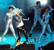 Singer with bright white hair on a stage wearing black leather jeans, coat and white shirt. Surrounded by 4 dancers wearing silver-grey body suits.