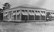 Photograph of a one-story building with shutters open and two men standing in front