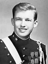A black-and-white photograph of son Donald Trump as a teenager, smiling and wearing a dark pseudo-military uniform with various badges and a light-colored stripe crossing his right shoulder