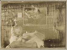 Old damaged and faded monochrome photograph of a woman wearing a pith helmet, long-sleeved and -skirted dress and apron. She is sitting beside a bed, over which hangs a religious picture and behind which is a wall made of reeds.