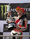A man in his mid-30s is wearing a red baseball cap and a multicoloured motorcycling overalls. He is holding a large silver trophy depicting a motorcycle in both his hands on a podium
