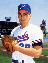 A man wearing a white baseball uniform with "Sounds" on the chest in blue and red and a blue cap with a white "N" on the center stands on a baseball field with his hands together in his brown leather glove.